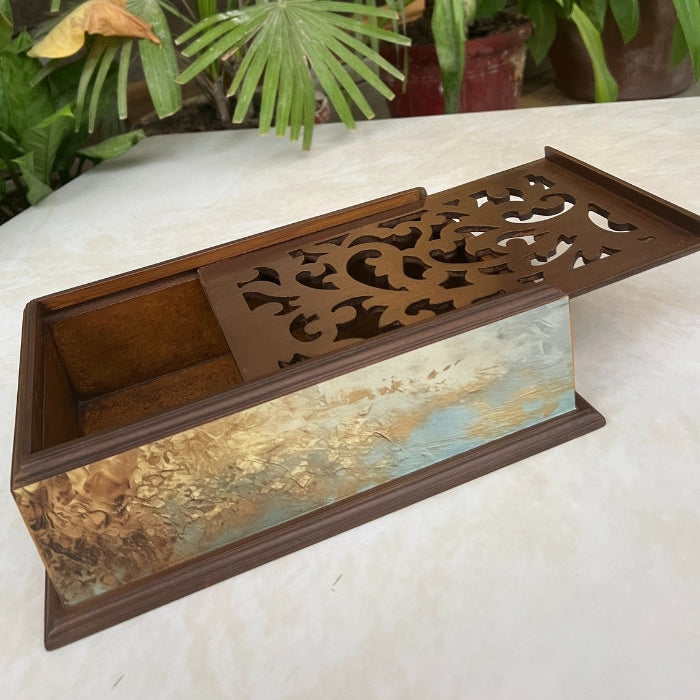 Decorative metal box with intricate design on a white surface with plants in the background
