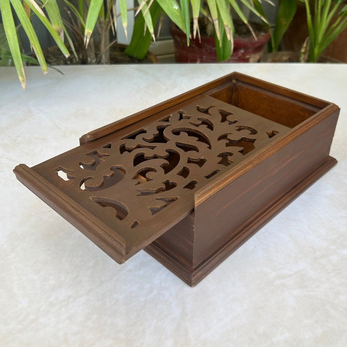 Decorative wooden box with intricate cut-out design on a white surface with plants in the background